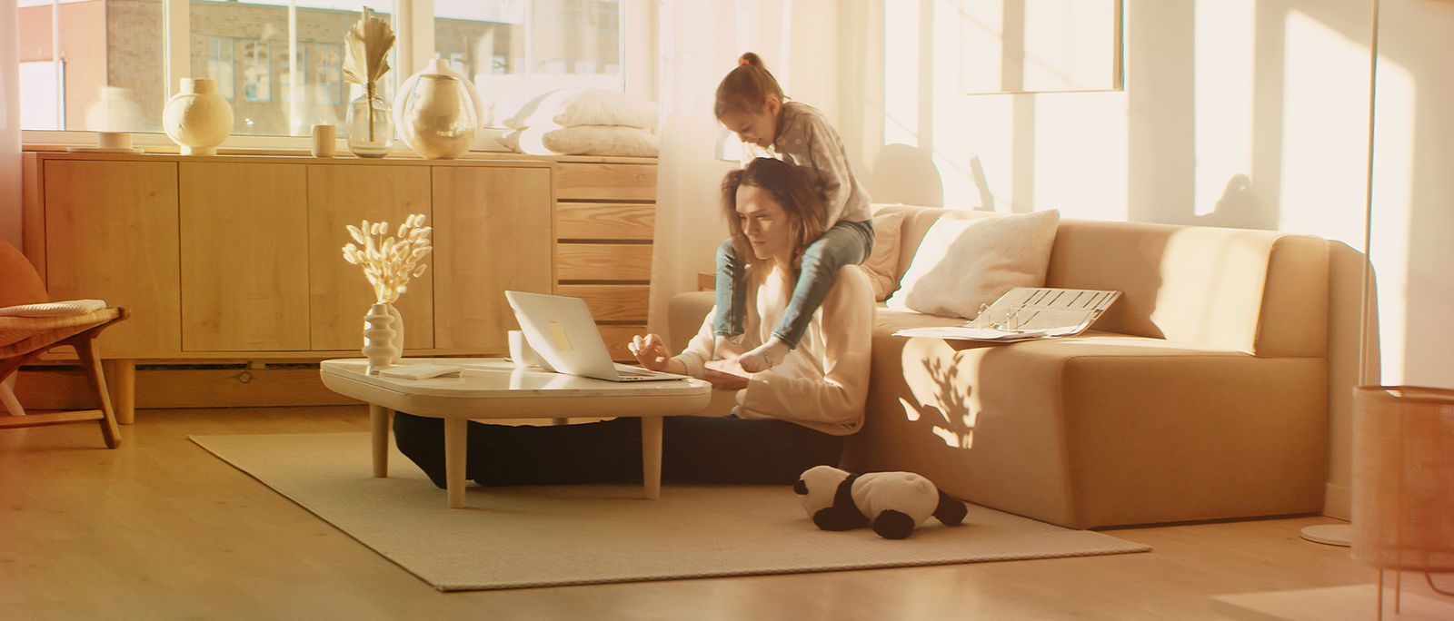 Mother sitting on the floor, with her laptop on the coffee table working from home while her daughter sits on her shoulders.